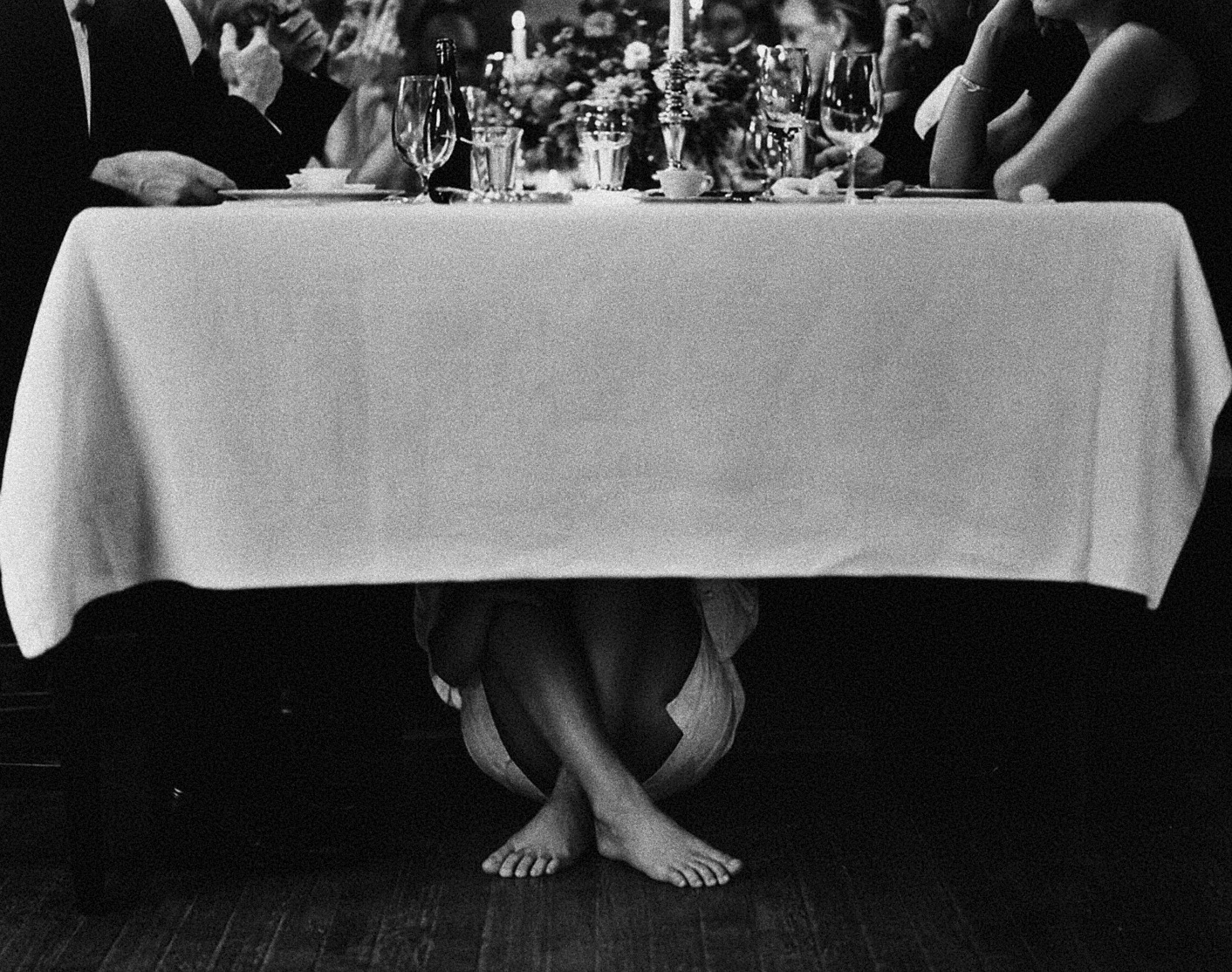 Black-and-white photograph of a dinner table, with a person's bare feet visible beneath the tablecloth.